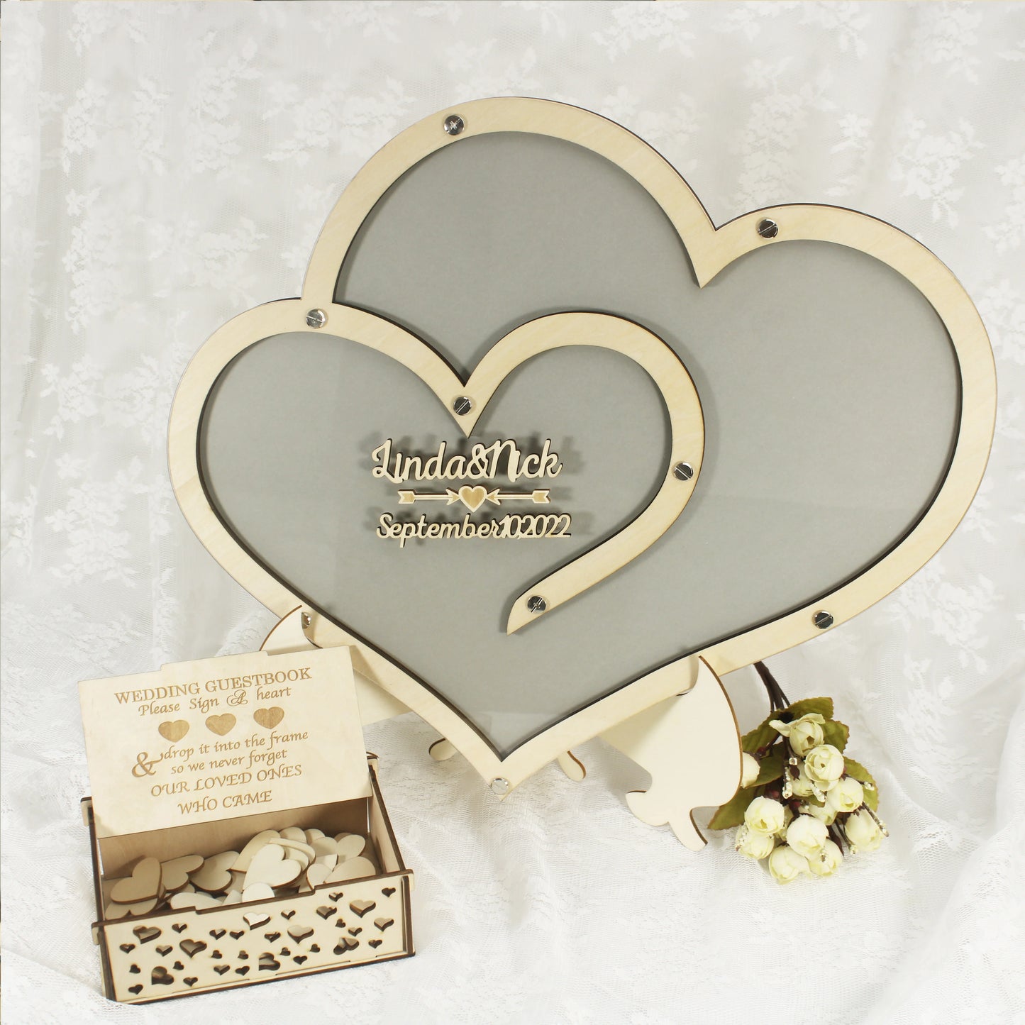 Wooden heart-shaped wedding guestbook frame with "Linda & Nick, September 10, 2022" engraved. A box with wooden hearts for signing is in front, alongside white roses.