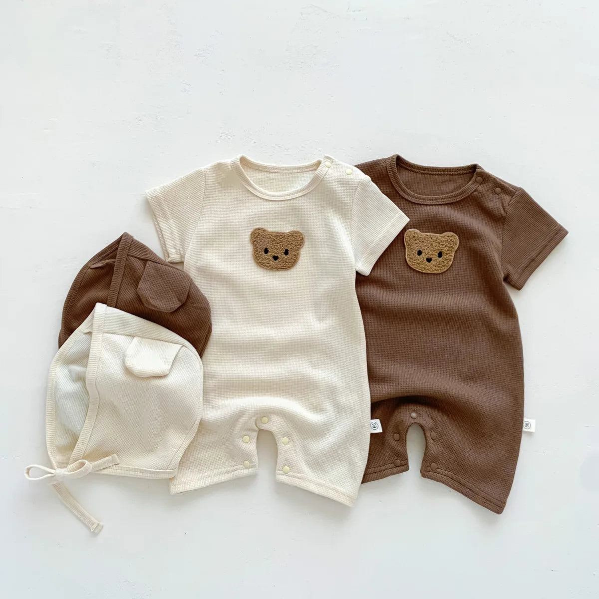Two baby onesies with bear faces, one cream, one brown, paired with matching bonnets. Cute, cozy outfits laid on a light background.