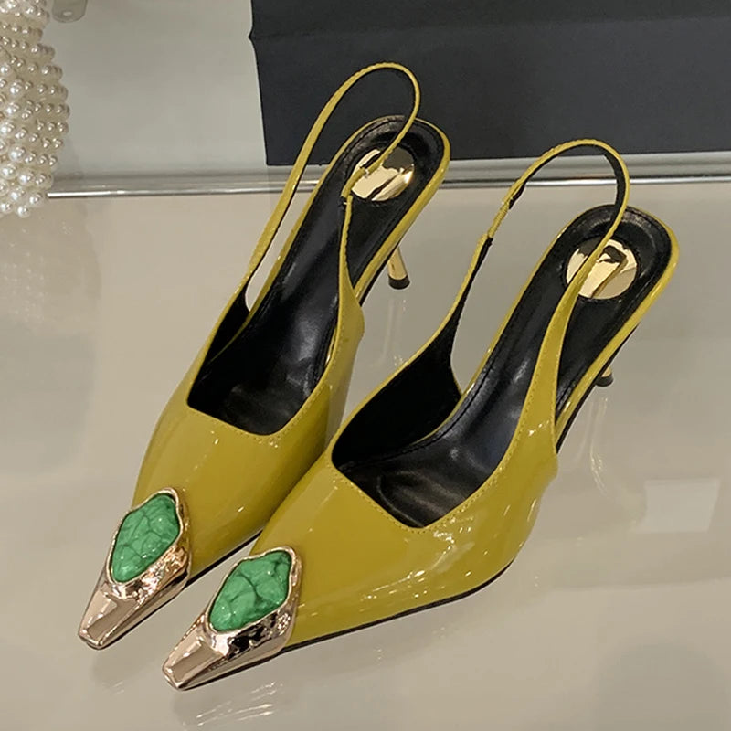 Yellow slingback heels with metallic gold accents and green stone embellishments on the pointed toes, displayed on a reflective surface.