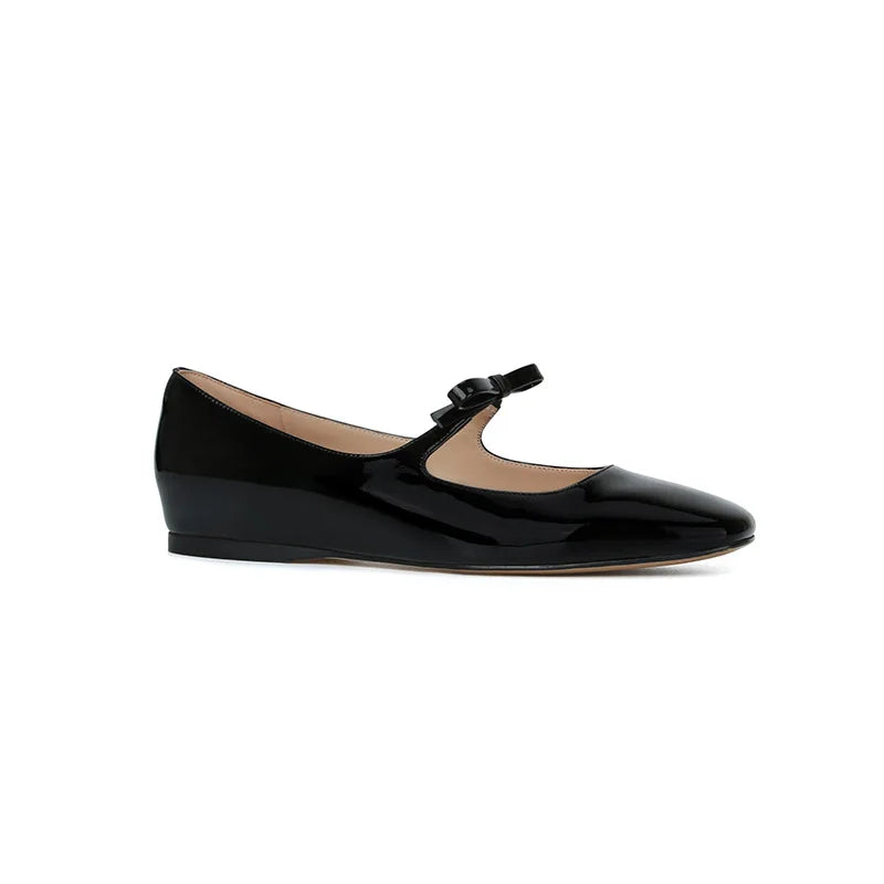 Black patent leather Mary Jane shoe with a wedge heel and a small bow on the strap, set against a white background.