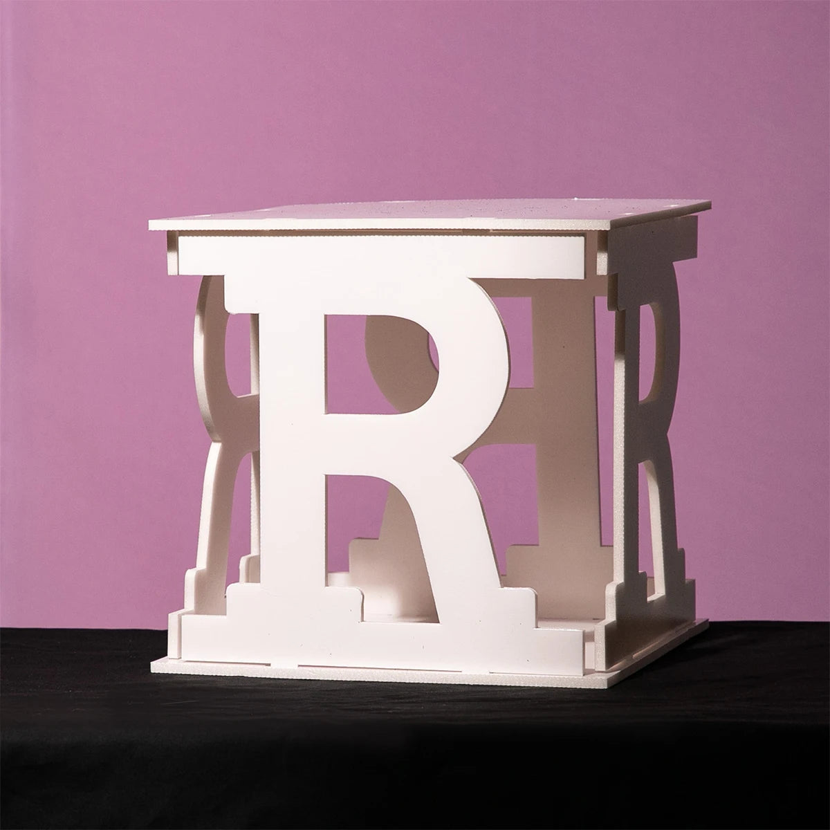 White cubic sculpture with large cut-out letters "R" on each face, standing on a black surface against a pink background.
