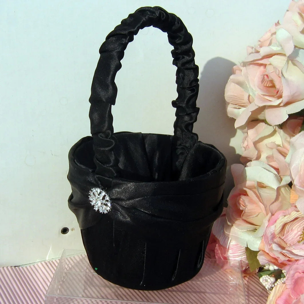 Elegant black satin basket with a decorative brooch sits on a striped surface next to a cluster of pink and white flowers, creating a stylish contrast.