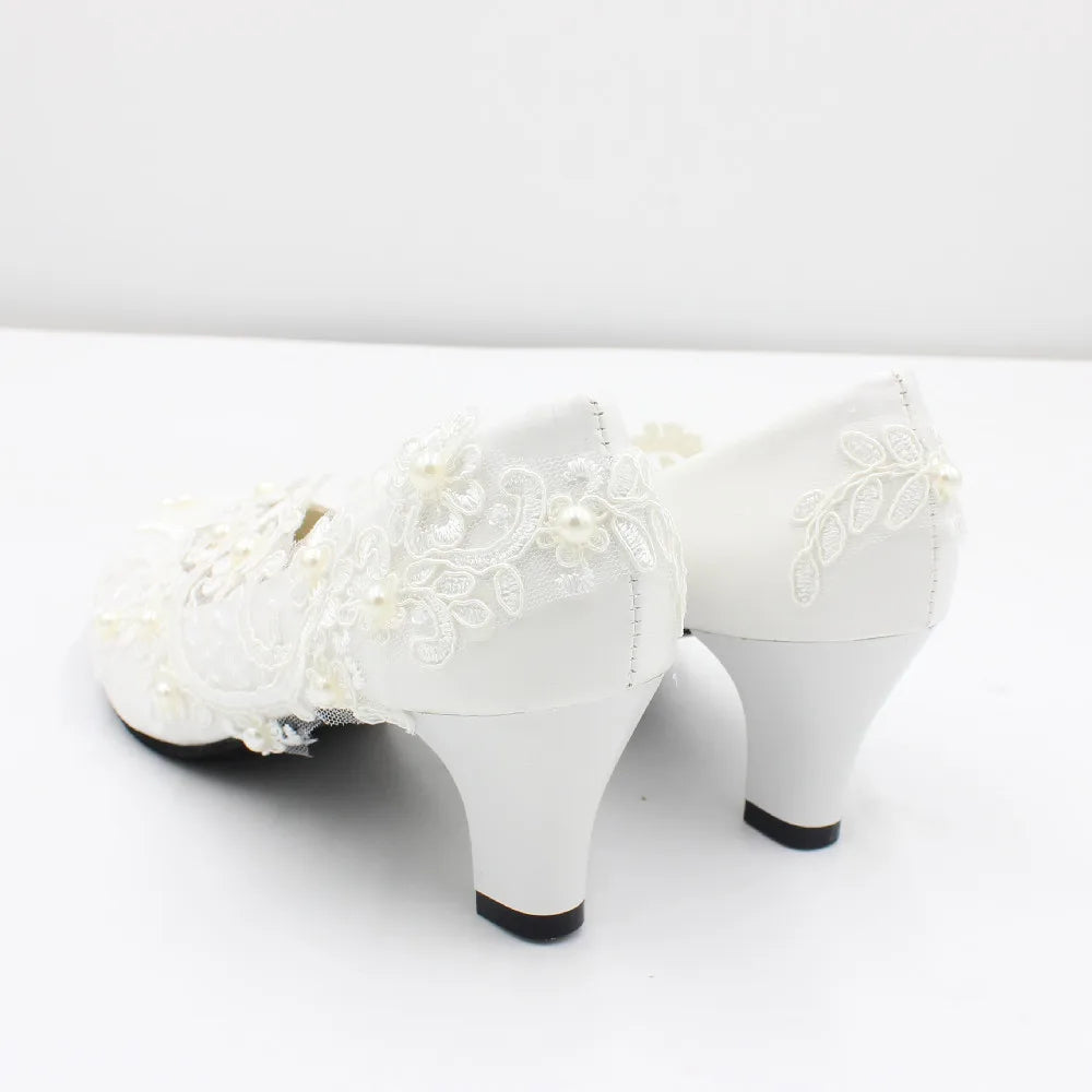 White satin bridal shoes with mid-height heels, adorned with delicate lace, floral embroidery, and pearl details, set against a plain white backdrop.