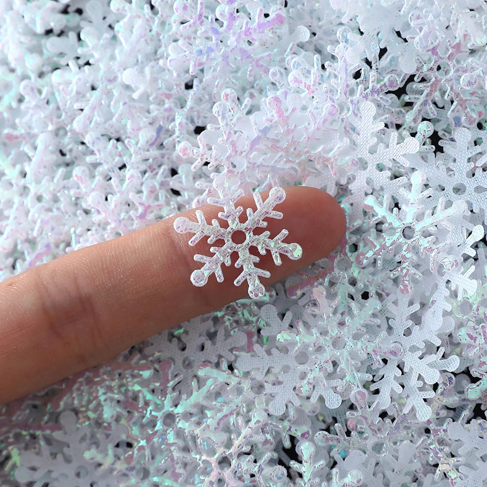 A fingertip holds a single translucent, glittery snowflake decoration among a pile of similar snowflakes.