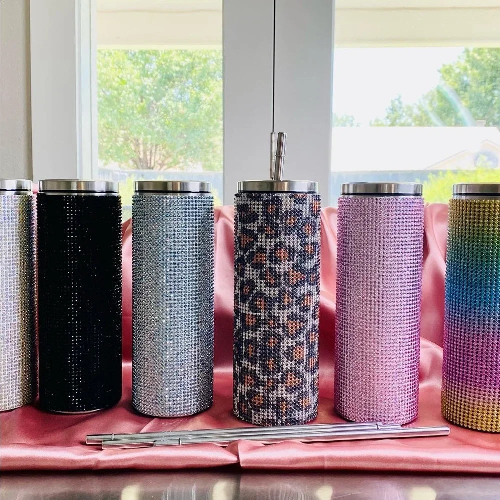 Array of colorful rhinestone-studded tumblers on a pink satin cloth, featuring leopard, ombre, and solid designs, with metal lids and straws. Bright window background.