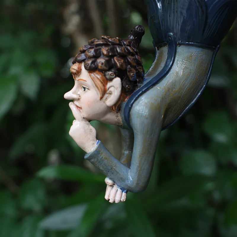 A whimsical garden sculpture of a pondering gnome with a pinecone hat, leaning on one hand. The figure is set against a blurred green foliage background.