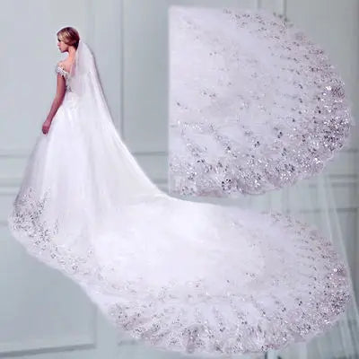 A bride in an elegant off-shoulder wedding gown stands with a long, ornate lace train adorned with intricate floral embroidery and shimmering details, set against a soft, neutral backdrop.
