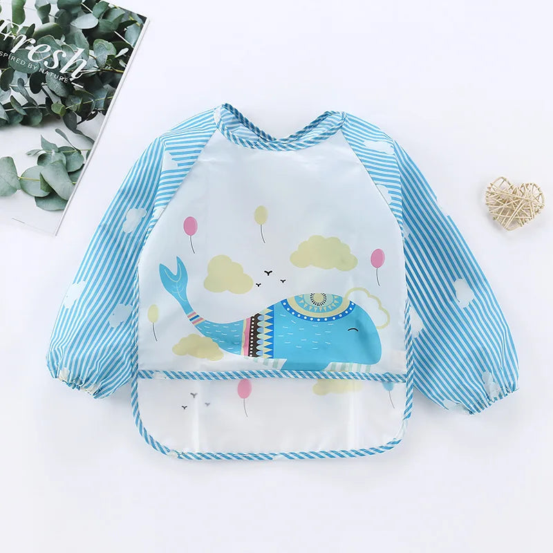 Blue waterproof bib with long sleeves featuring a smiling whale, clouds, and balloons design. Striped sleeves and hem. A plant and a woven heart decor are in the background.