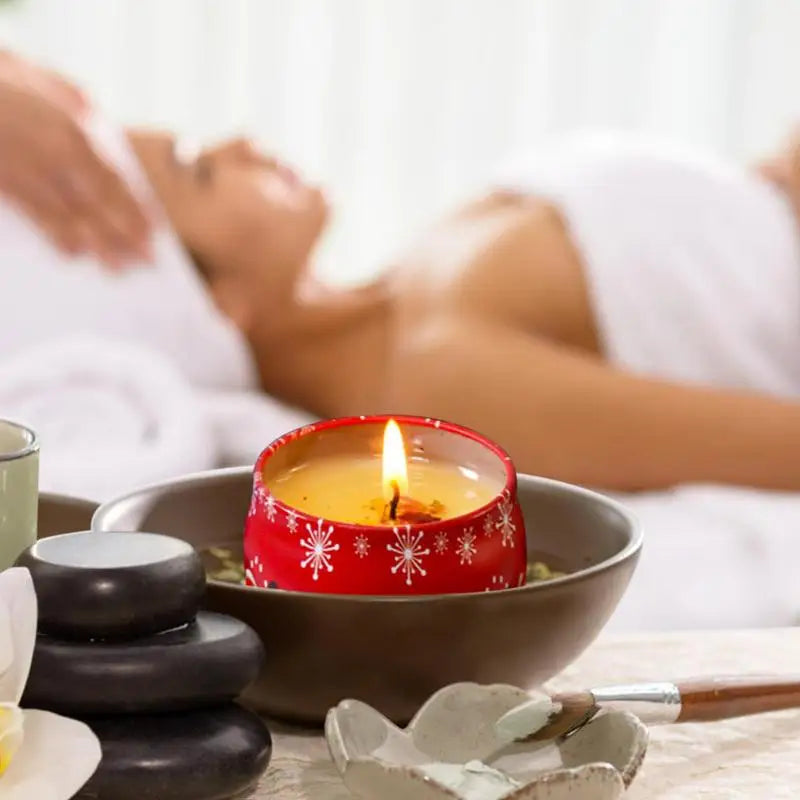 Red candle burning in a bowl next to stacked stones and spa items. In the background, a person lies on a massage table, covered with a towel, suggesting a relaxing spa setting.