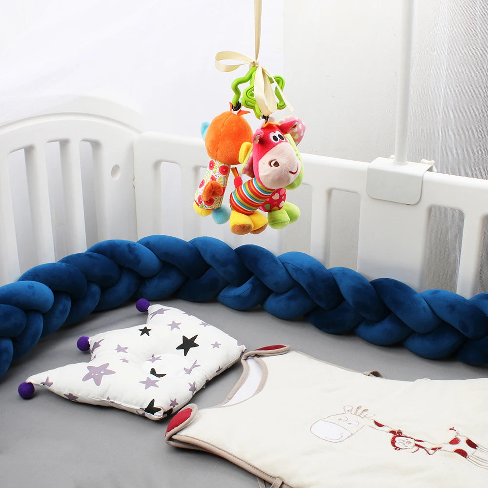Baby crib with a blue braided bumper, hanging colorful plush toys, a star-patterned pillow, and a baby sleeping bag featuring a cute animal design.