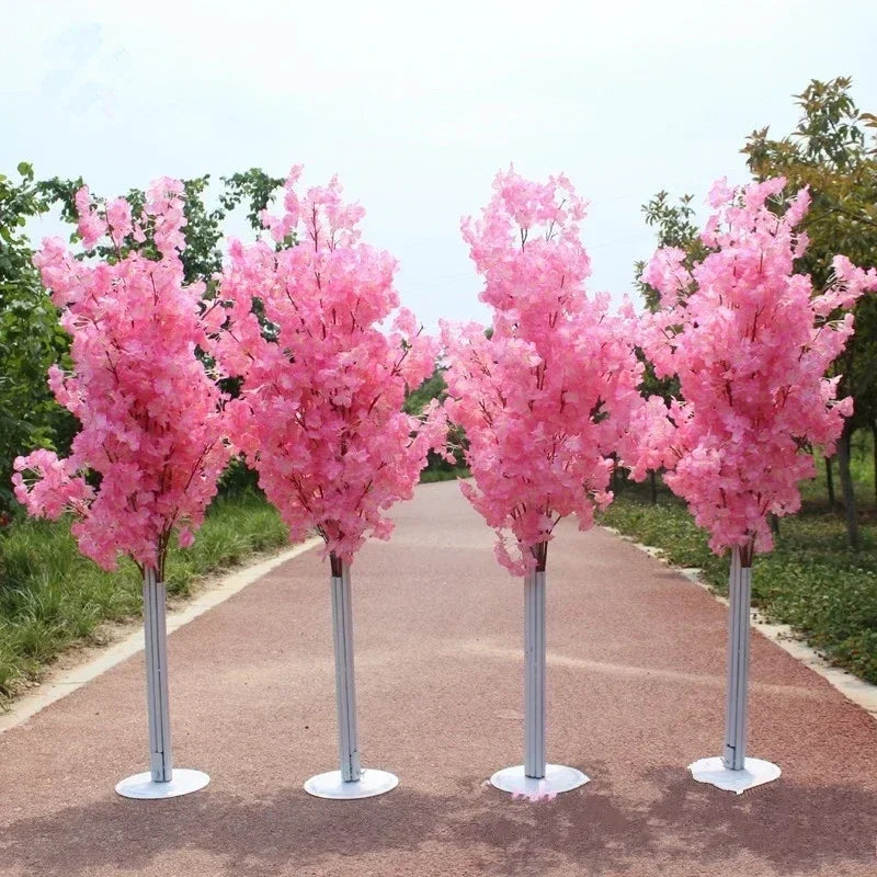 Four artificial cherry blossom trees with vibrant pink flowers stand on a paved path surrounded by greenery, under a clear sky.