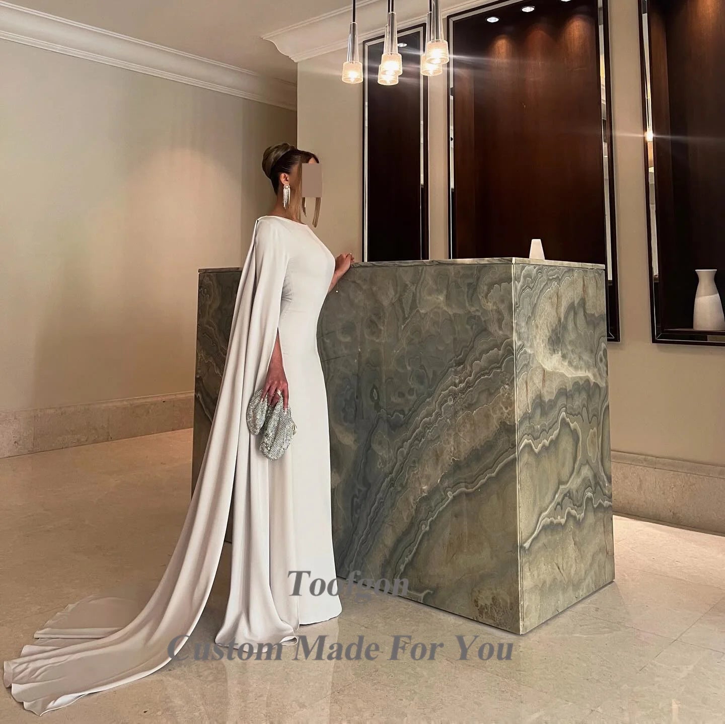 Elegant woman in a long white gown with a train and cape standing by a marble counter. She holds a sparkling clutch under chic pendant lights in a modern setting.