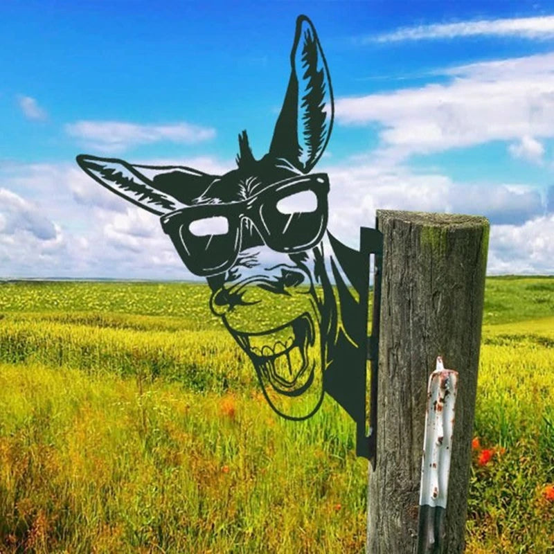 Metal art of a laughing donkey wearing sunglasses attached to a wooden post in a green field under a blue sky with clouds.