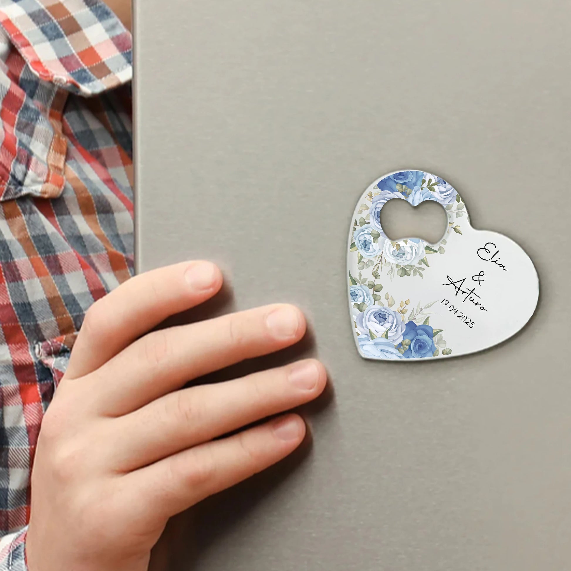 A person wearing a plaid shirt holds a grey surface with a heart-shaped magnet. The magnet is decorated with blue flowers and text, appearing to commemorate a special date.