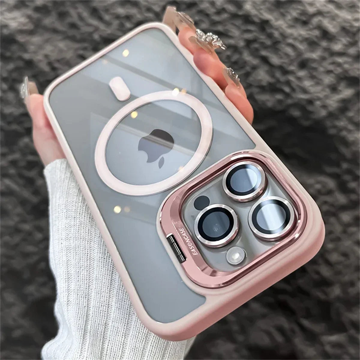A hand with manicured nails holds a smartphone with a pink and gray case featuring a circular pattern and dual rear camera lenses. The phone is against a textured dark surface.