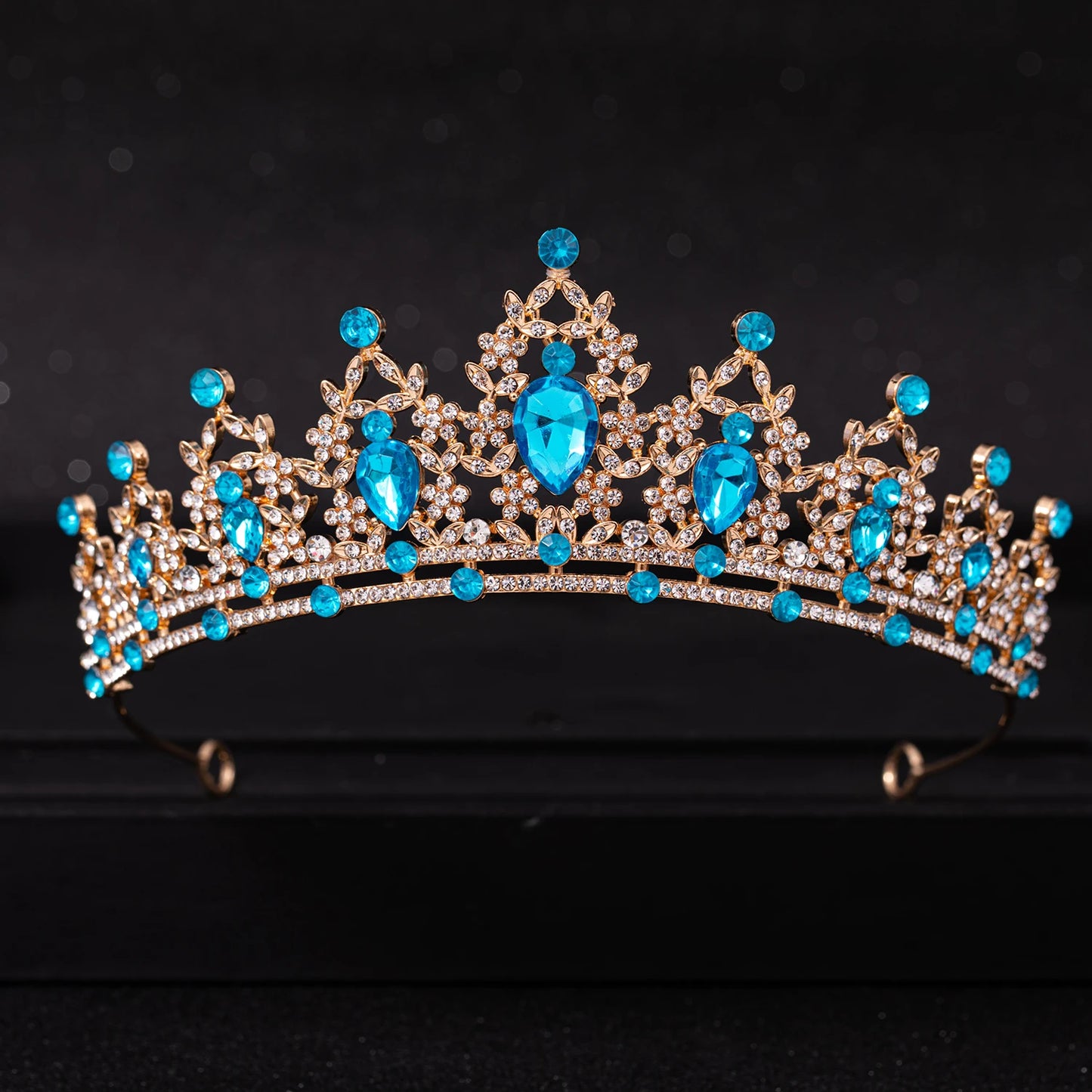 A gold tiara adorned with large teardrop-shaped blue gems and clusters of smaller white crystals, set against a black background.