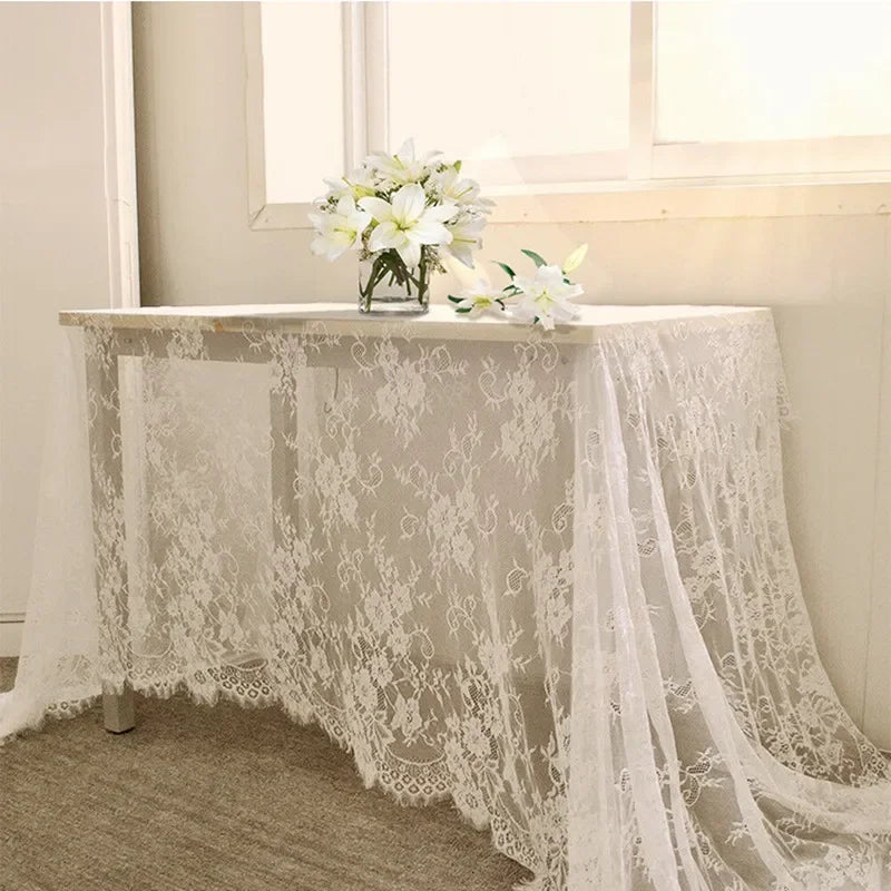A table draped with a white lace cloth, topped with a vase of white flowers, is positioned against a wall. Soft sunlight illuminates the scene, creating a serene atmosphere.