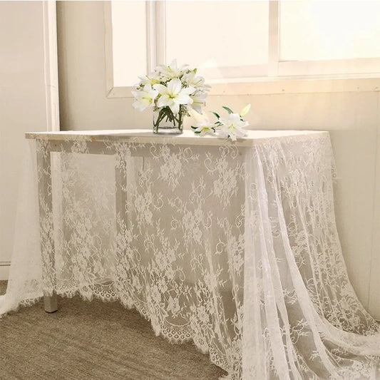 A table draped with a white lace cloth, topped with a vase of white flowers, is positioned against a wall. Soft sunlight illuminates the scene, creating a serene atmosphere.