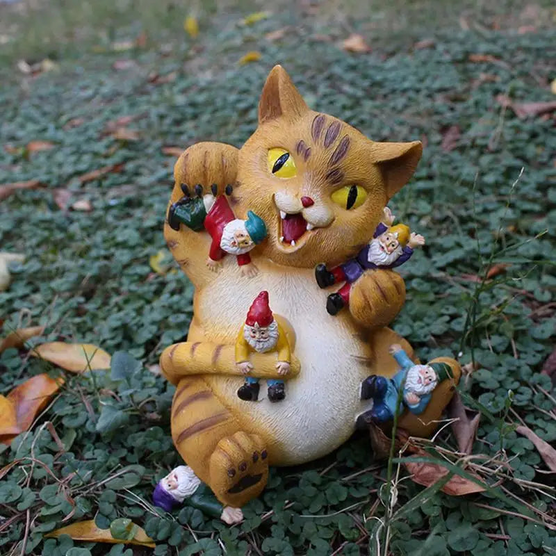 A playful garden statue of a smiling cat surrounded by tiny gnomes clinging to its paws and body, set on green grass with scattered leaves.