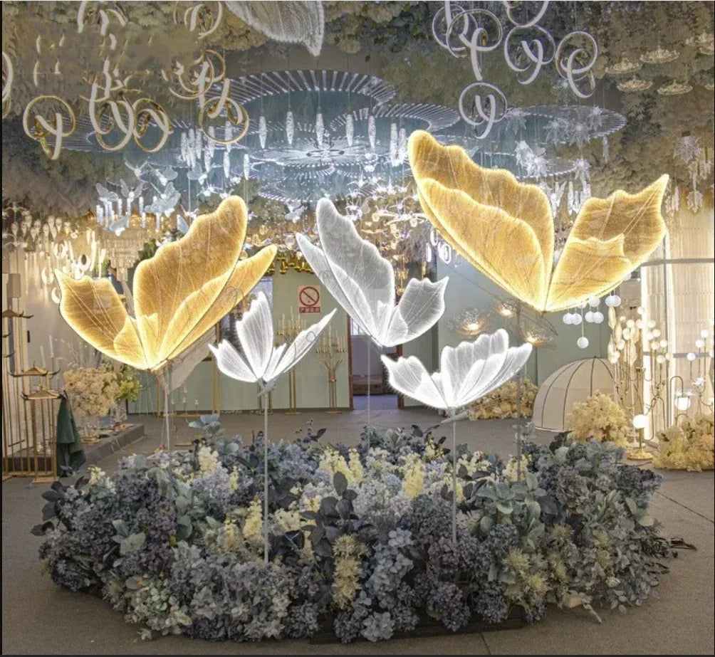 Illuminated butterfly wings float above a lush floral display in an ornate room with chandeliers and intricate ceiling designs, creating a whimsical, magical atmosphere.