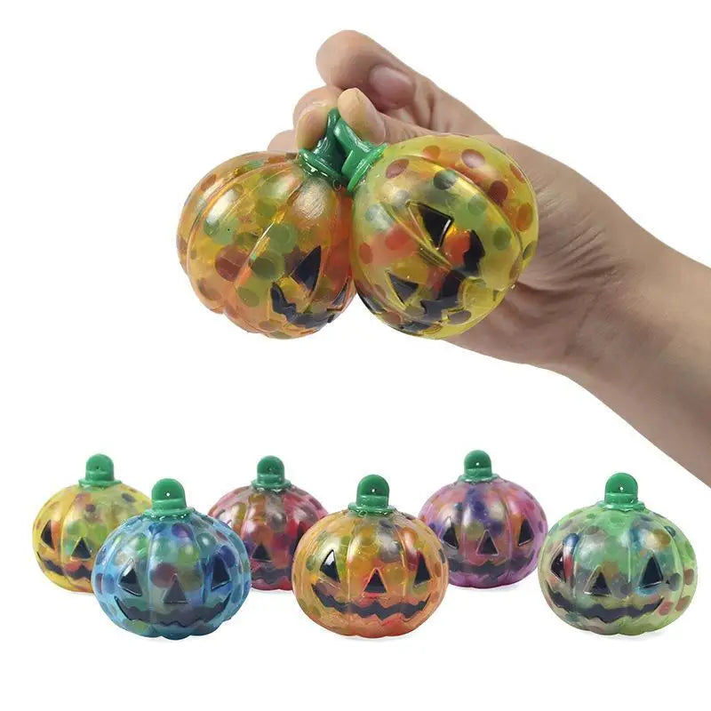 A hand squeezes two transparent pumpkin stress balls filled with colorful beads. Below, five more pumpkins in various colors rest on a white surface, each with a jack-o'-lantern face.