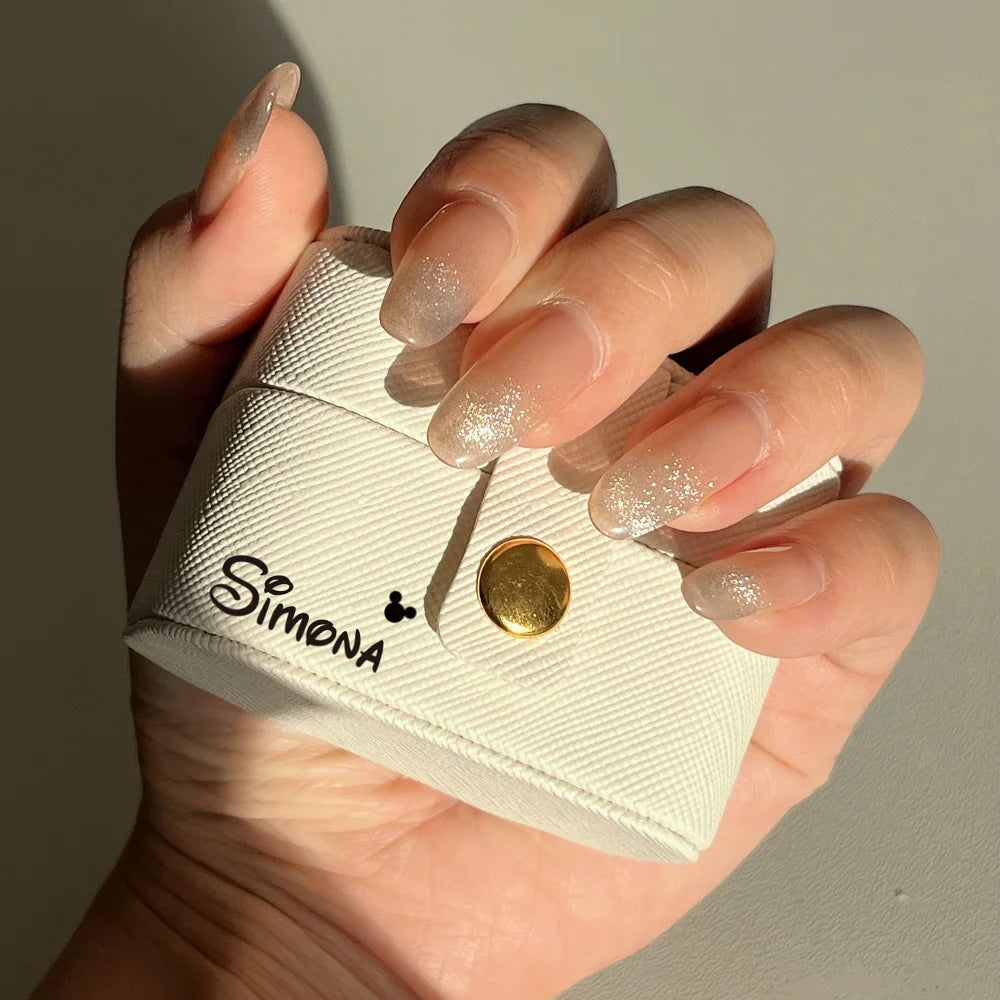 Hand with glittery, translucent nails holding a white textured case with "Simona" and a small design printed on it, featuring a gold button closure.