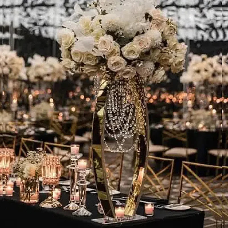 Elegant wedding table setting featuring a tall floral centerpiece with white roses and hanging crystals, surrounded by numerous lit candles and gold-accented chairs.