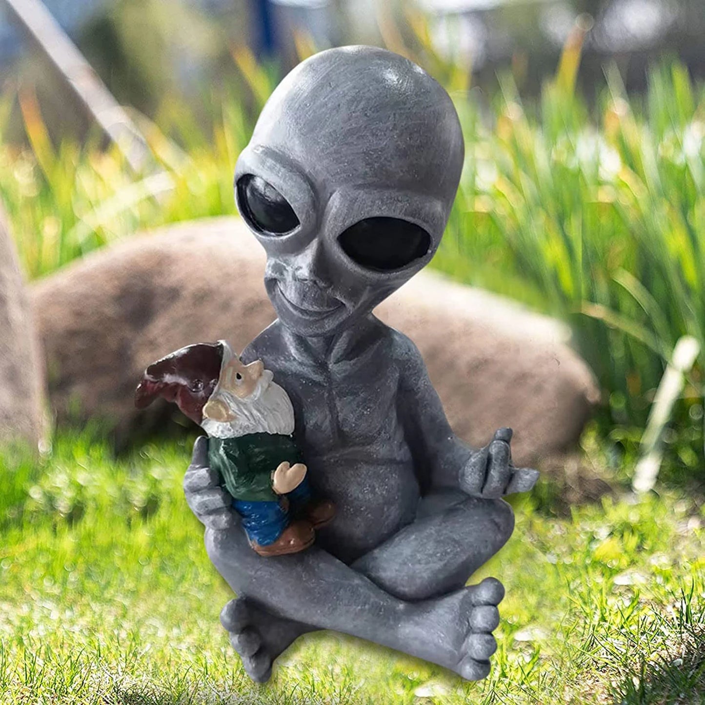 A gray alien statue sitting cross-legged on grass, holding a small garden gnome. The scene is outdoors with blurred greenery and a rock in the background, conveying a whimsical feel.