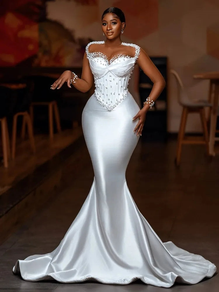 A woman in a white, pearl-embellished mermaid gown poses confidently indoors. The dress features a fitted bodice with intricate beading and a flowing skirt with a small train.