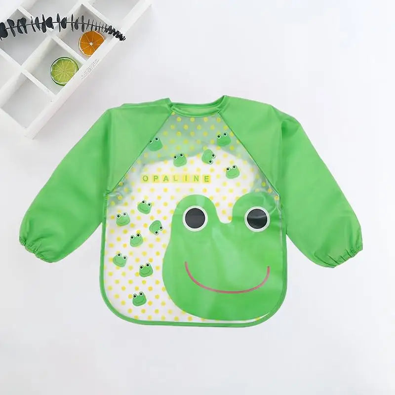 Green children's apron with long sleeves, featuring a frog face and pattern on a yellow polka-dot background. Shelf with citrus slices and decorative branch in the corner.