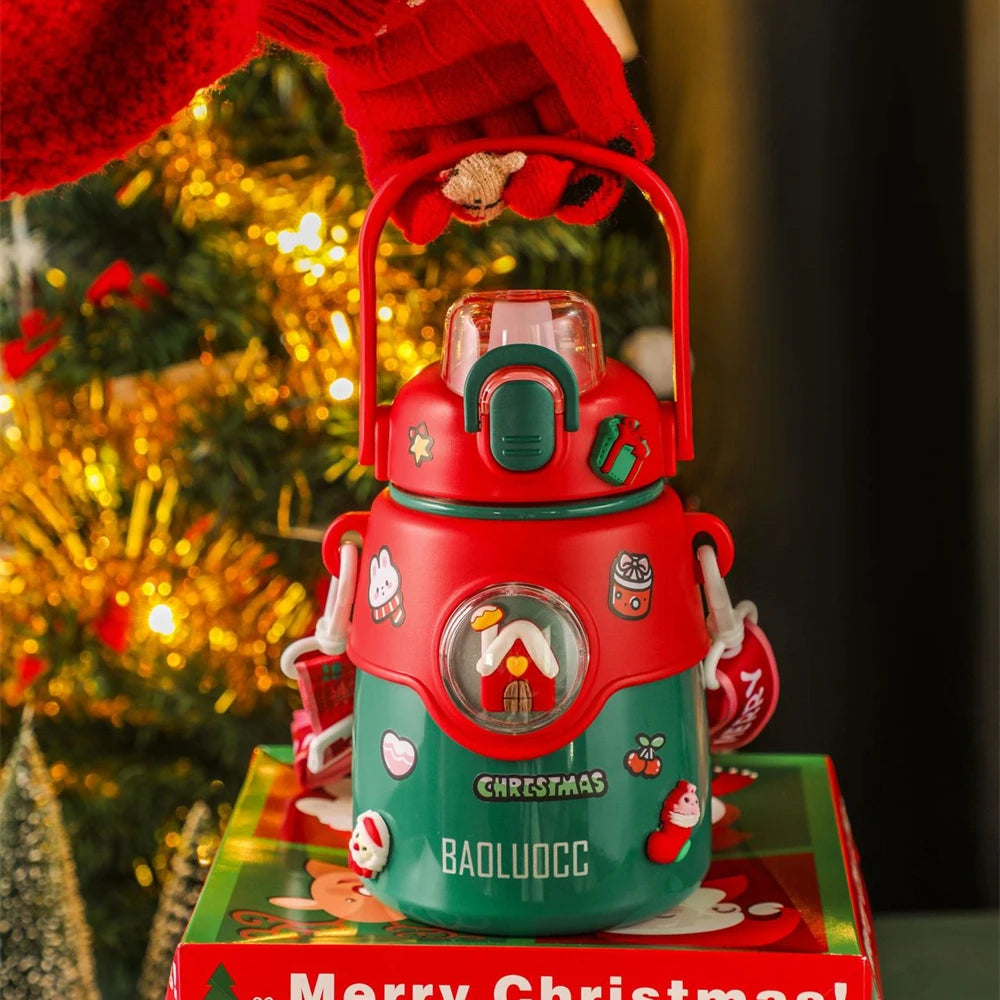 Red and green festive thermos with Christmas decorations, held by a hand in a red glove. A Christmas tree with lights is in the background, and a "Merry Christmas" box is underneath.