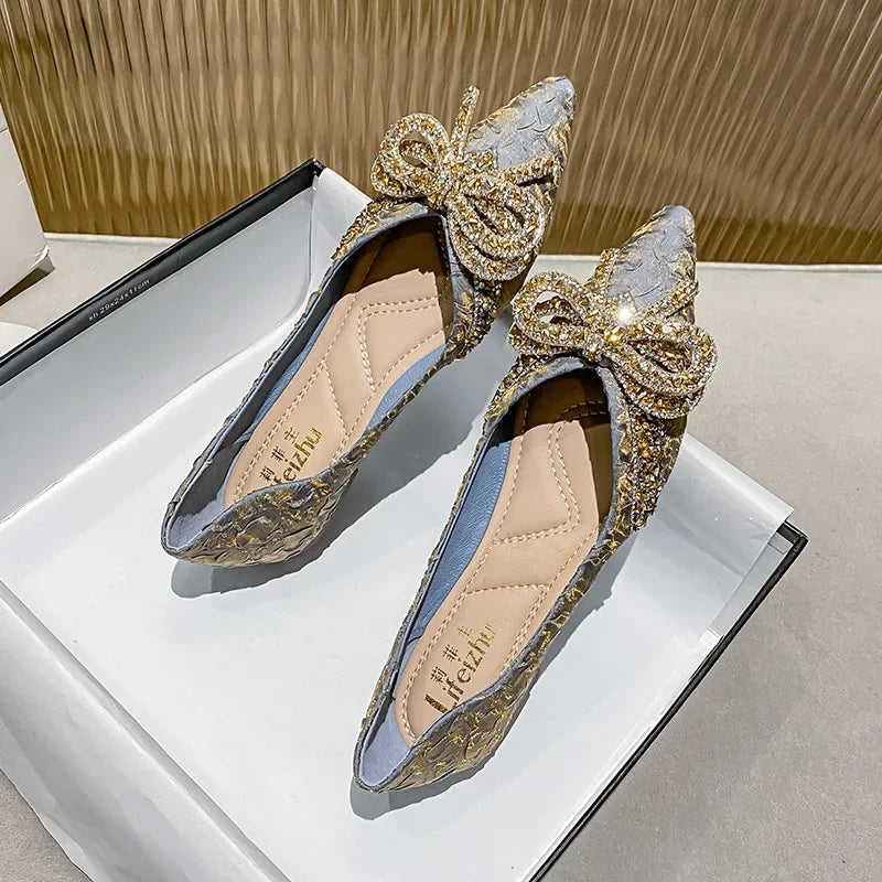 Elegant pointed-toe flats with intricate golden embroidery and large decorative bows, displayed in an open white shoe box on a textured background.