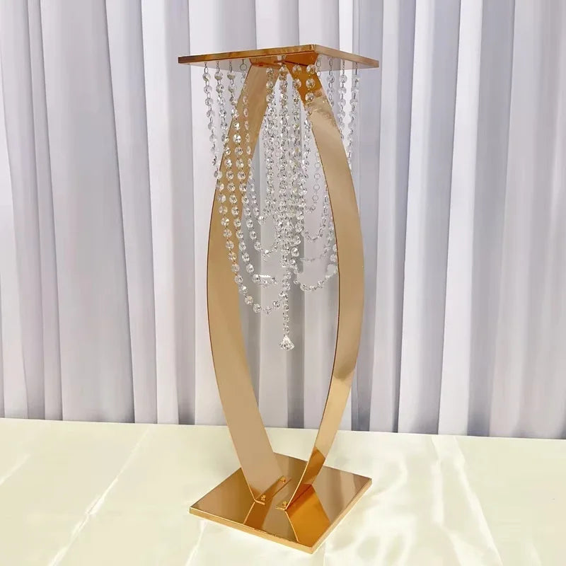 Gold decorative stand with a square base and top, featuring draped crystal strands. Set against a white draped curtain background, placed on a light fabric surface.