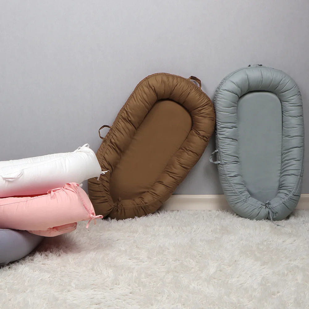 Baby nests in white, pink, brown, and blue are arranged on a soft white carpet against a light gray wall. Two nests are leaning against the wall; the others are stacked.