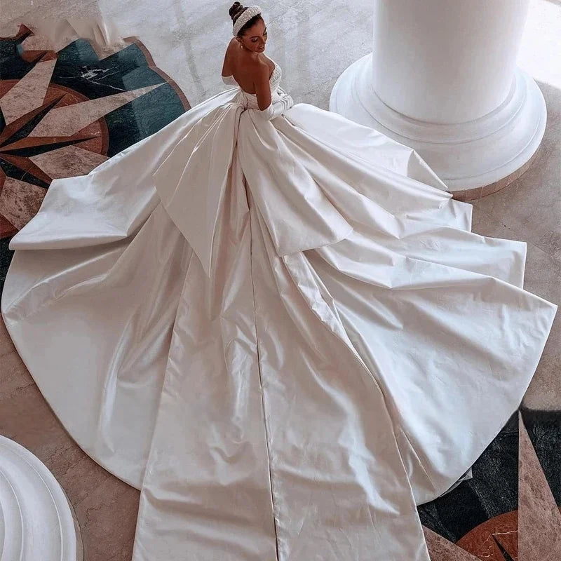 Bride in an elegant white satin gown with a large train, standing by a pillar. The dress has a bow at the back and the setting features a decorative marble floor.