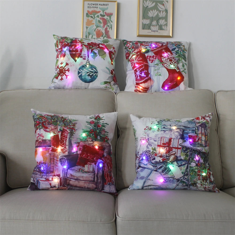 Four festive pillows with Christmas designs and colorful LED lights are arranged on a beige sofa. Above are two floral prints framed on the wall.