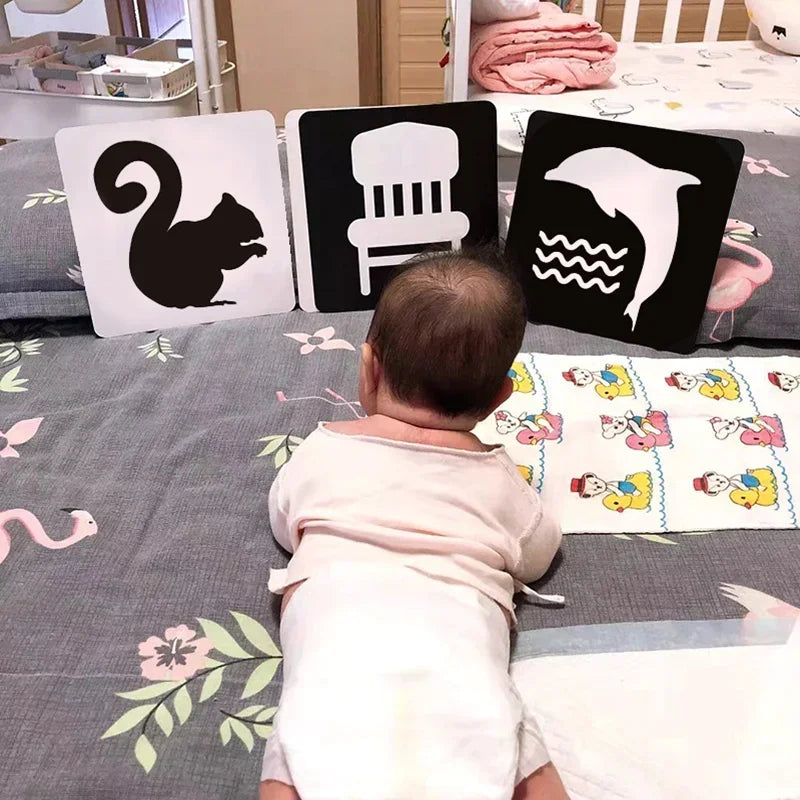 A baby lying on a bed looks at three high-contrast cards showing silhouettes of a squirrel, chair, and dolphin. The bed has a floral pattern and cartoon illustrations.
