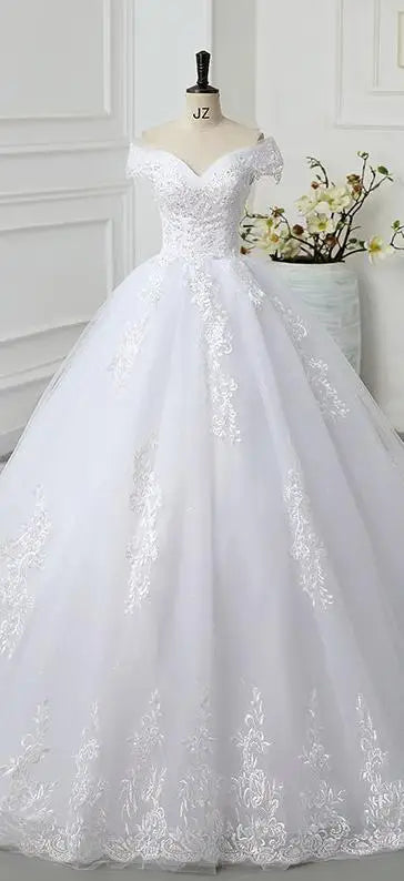 Elegant white wedding gown on mannequin with lace bodice, off-shoulder sleeves, and voluminous tulle skirt. Soft floral embroidery details. White walls and flowers in background.