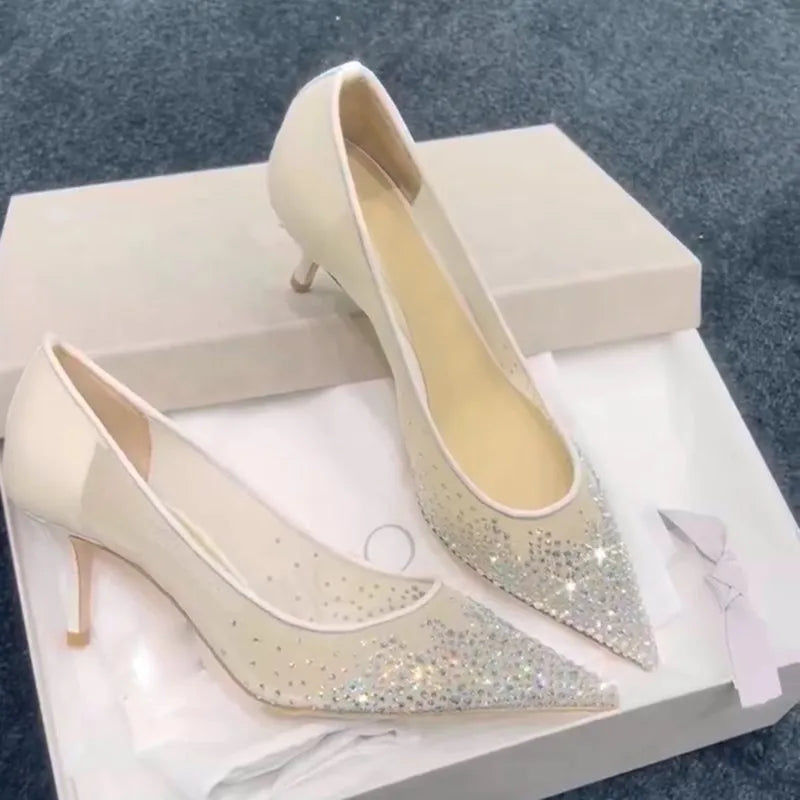 Elegant beige high heels with sparkling rhinestones on pointed toes, displayed in an open shoebox against a soft fabric background.