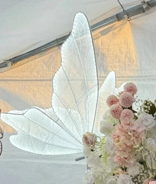 Delicate white fairy wings are displayed against a tent backdrop, accompanied by a bouquet of pink and white flowers, creating a whimsical and ethereal scene.