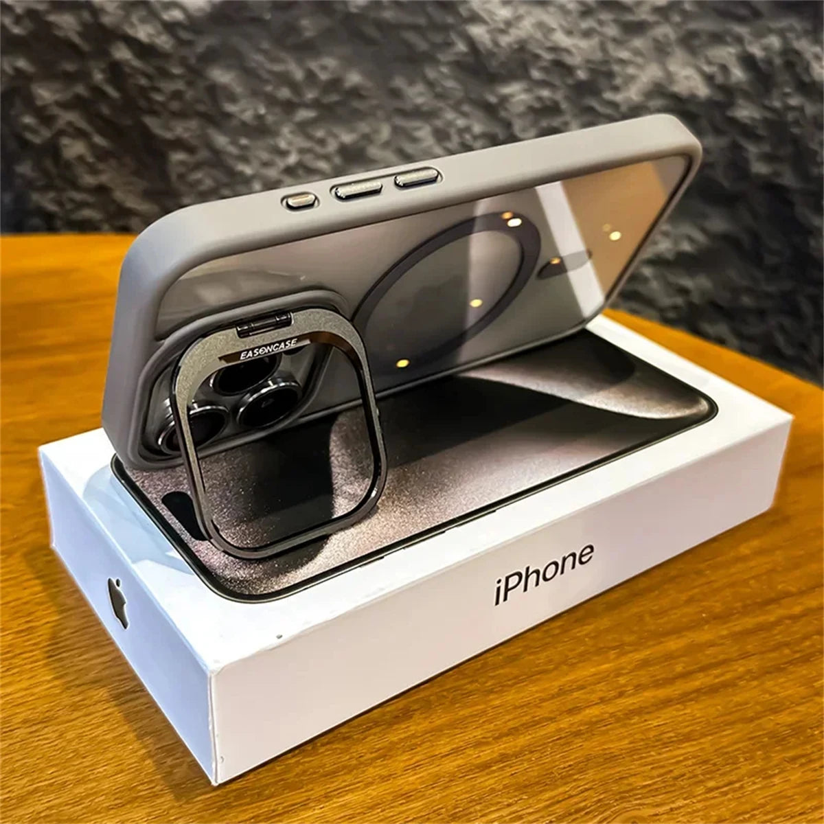 Gray smartphone case with a foldable ring stand on an iPhone box, placed on a wooden surface. The phone has a triple-lens camera and a MagSafe-compatible design.