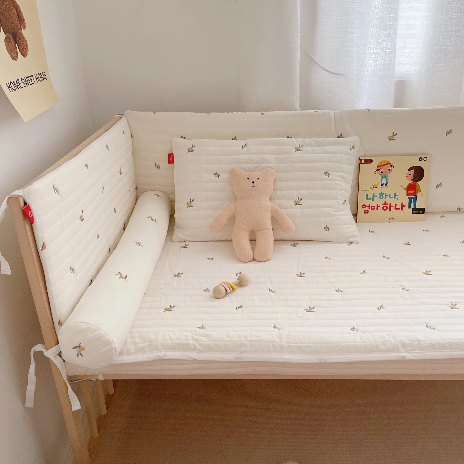 Cozy crib with white bedding featuring small leaf designs, a beige teddy, a colorful rattle, and a children's book. A "Home Sweet Home" poster hangs on the wall.