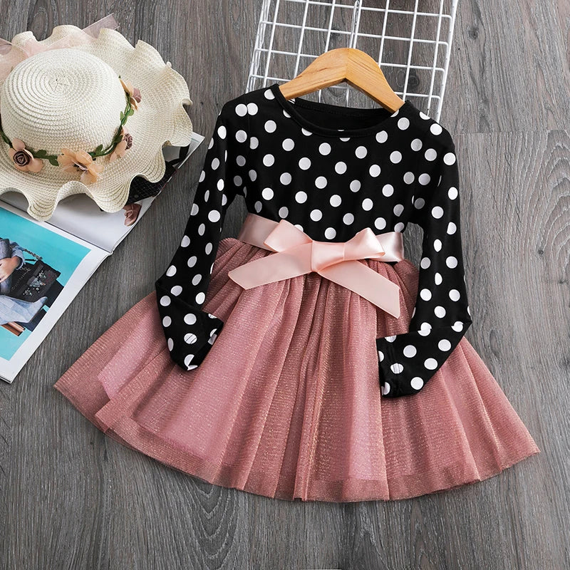 Polka dot top with long sleeves and a pink bow over a layered pink tulle skirt, styled on a hanger beside a floral sun hat on a gray wood floor.