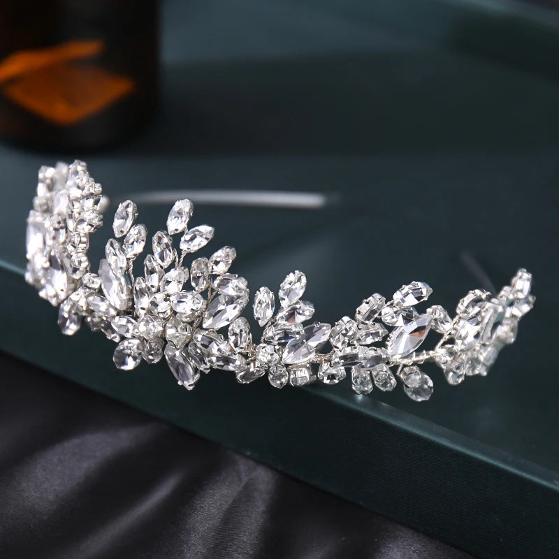 A sparkling silver tiara adorned with clear crystals in various shapes, set against a dark green background and black surface.