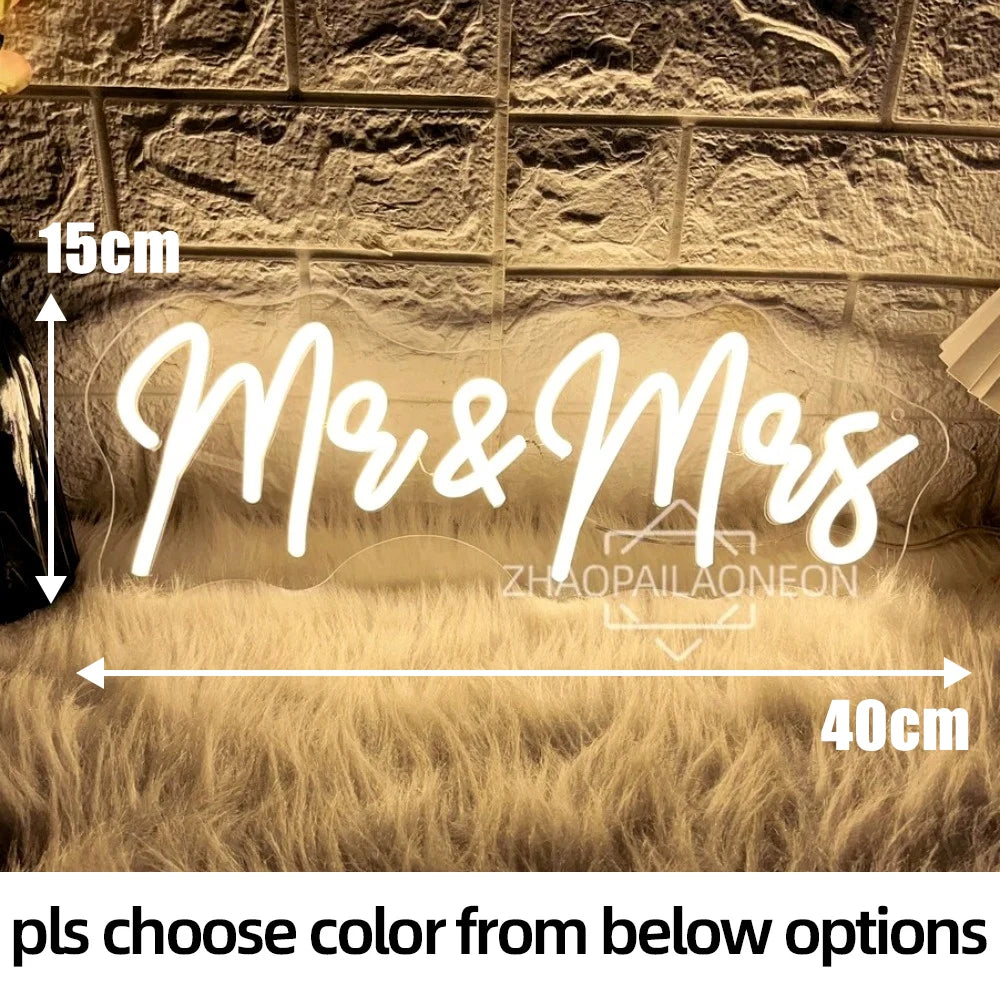 Neon sign reading "Mr & Mrs" with dimensions 15cm by 40cm, illuminated in warm light on a textured brick wall background and a fluffy surface. Text reads: "pls choose color from below options."