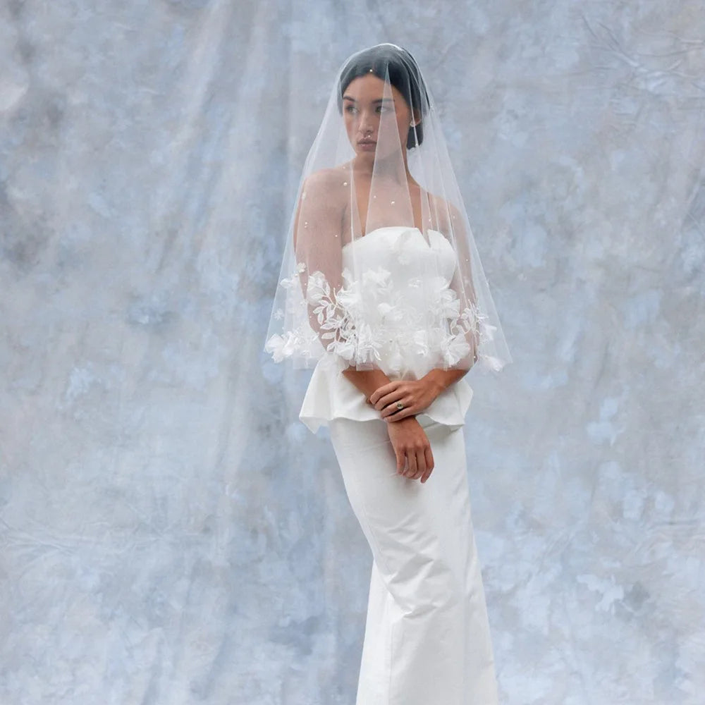 Bride in a strapless white gown with floral lace, wearing a veil with pearl accents. She stands against a soft blue-gray mottled backdrop, hands clasped in front.