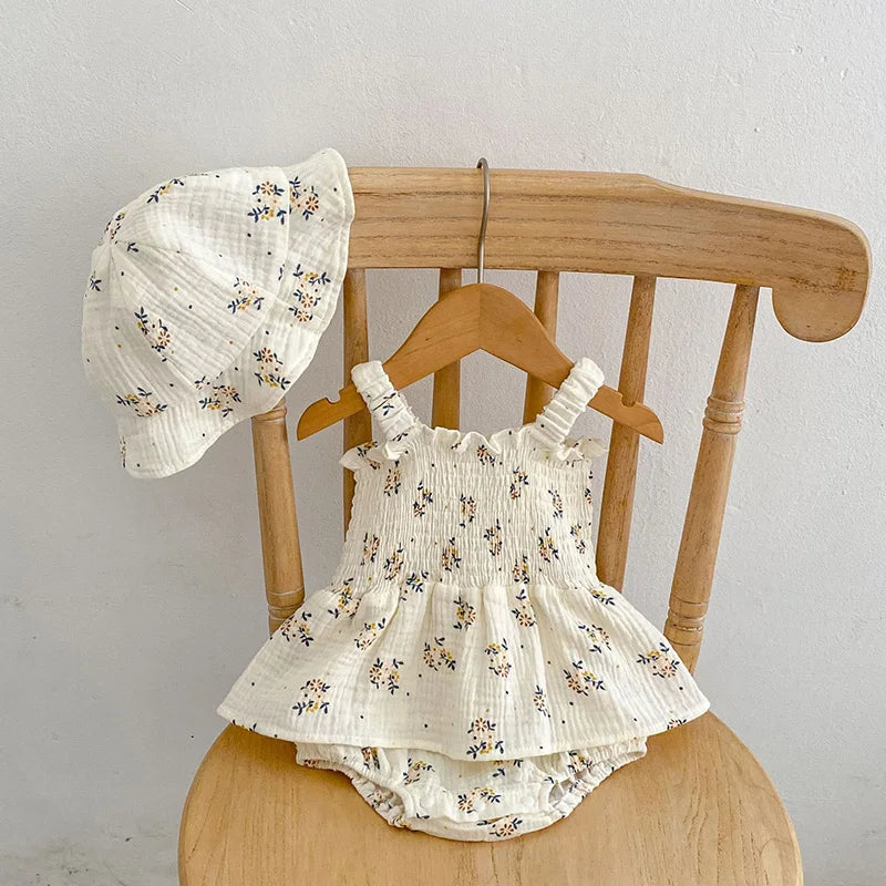 A light floral baby outfit with a ruffled dress, bloomers, and matching hat is displayed on a wooden chair. The fabric is cream with small, delicate flowers.