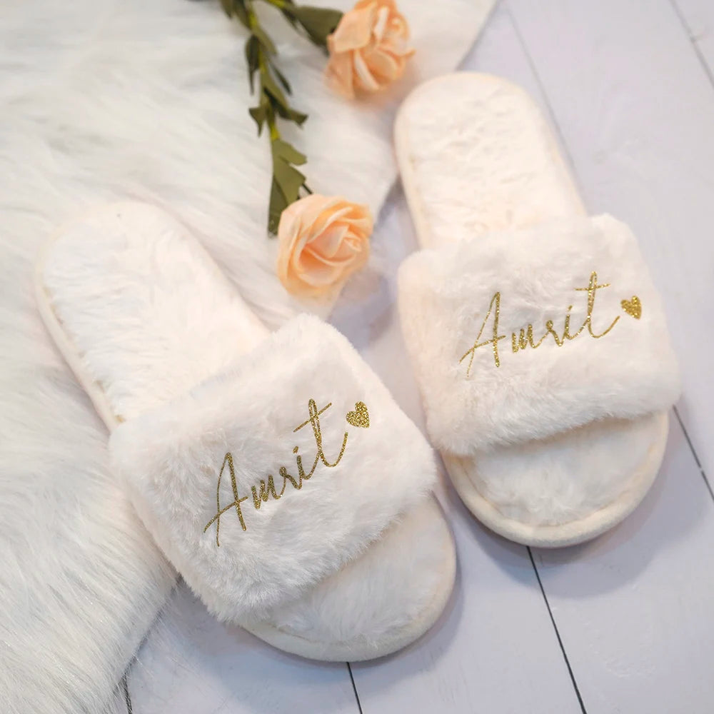 White fluffy slippers with gold "Amrit" text and heart detail. Placed on a soft white surface with two light orange roses for a cozy and elegant look.