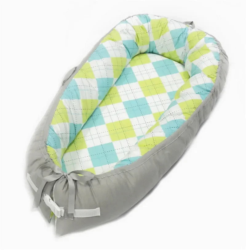 Cozy oval baby nest with soft gray sides and a colorful green, blue, and white argyle pattern inside. Features a bow tie detail and adjustable straps for secure fit.