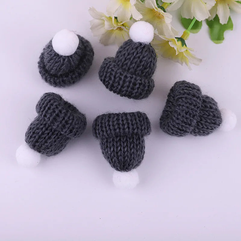 Five miniature gray knitted hats with white pom-poms arranged on a white background, next to a cluster of light yellow flowers with green leaves.